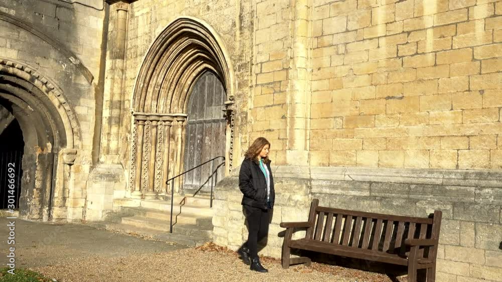 A pretty young woman walking casually in the autumn sunshine past a side door then sitting down on a wooden bench next to the ancient and historic cathedral, in the city of Peterborough, England, UK.