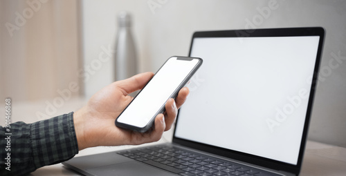 Close-up of male hand holding smartphone over laptop with mockup, white blank screen.