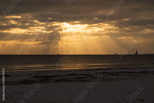 Sailboats on the horizon as the sun peeks through clouds with beams of light during sunset on Fort Myers Beach