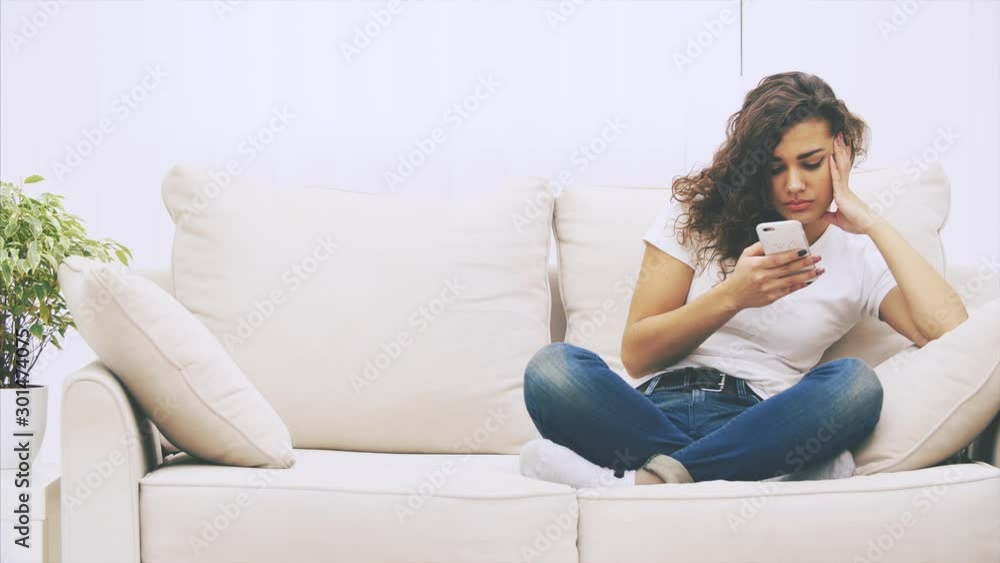 Lovely kinky afro-american girl is sitting on the sofa in lotos position, with phone in her hands, messaging. Her friend joining her. They start talking. Lovely kinky afro-american girl is sitting on the sofa in lotos position, with phone in her hands, messaging. Her friend joining her. They start talking.