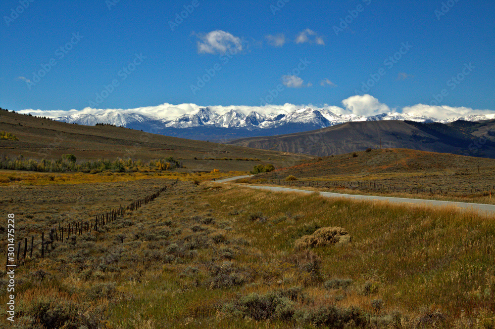 Highway threads its way thru the fall colors of a mountain valley with snow covered peaks in the background.