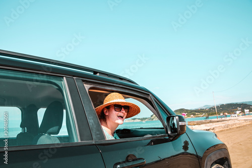 Young woman in a straw hat and sunglasses. Dark soul car on the beach. View of beautiful sea and blue sky. Place for your text or product. A beautiful summer day. Car trip and sightseeing of the coast