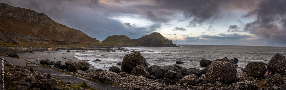 Panoramic view of the pathways and the bay at the coast called Giant's Causeway, a landmark in Northern Ireland, with the cliffs that surround the place and dramatic cloudy sky. Panoramic view of the pathways and the bay at the coast called Giant's Causeway, a landmark in Northern Ireland, with the cliffs that surround the place and dramatic cloudy sky.