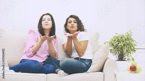 Beautiful different young girls sitting on the sofa side to side, posing, giving airkiss to the camera. photo