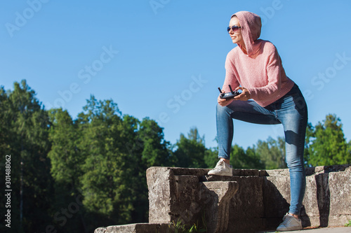 Young woman wearing sunglasses and hood on his head holding a remote control, carefully looks into the distance against.
