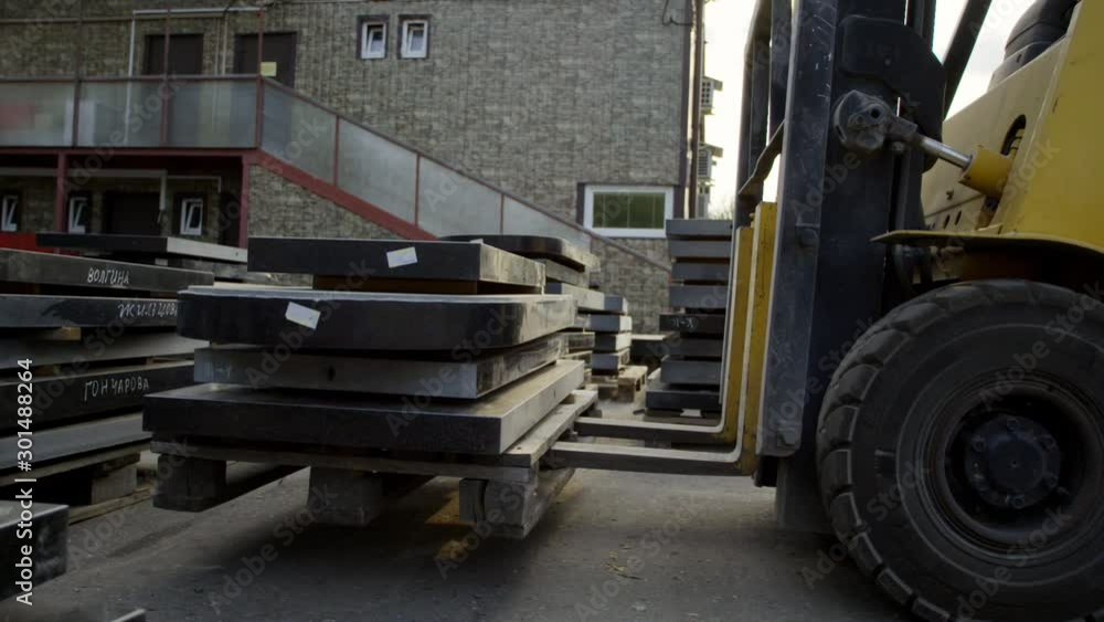 View of yellow forklift pallets lifter heavily loaded with dark granite blocks is lifting them up at outside area of some warehouse. View of yellow forklift pallets lifter heavily loaded with dark granite blocks is lifting them up at outside area of some warehouse.