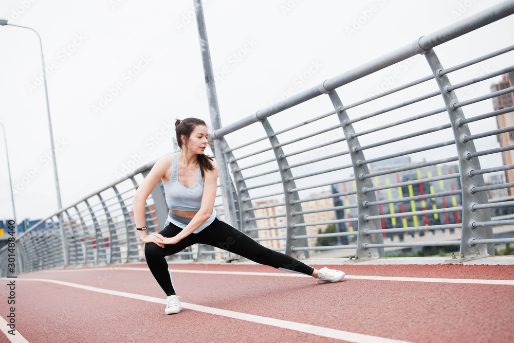 City fitness. A young sports woman in fitness suit is warming up, doing stretching against steel railing of the bridge.