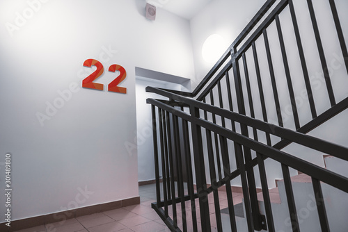 A typical flight of stairs in an office business skyscraper: clean white walls, metal fencing of the stairway, bright light, door to the floor, big orange digits "22", and the fire alarm speaker above