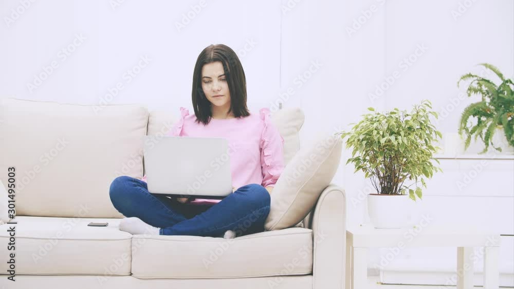Cute caucasian brunette girl sitting on the sofa in lotos position, with laptop, surfing in the internet, throwing hands, raising eyebrows, looking confused. Cute caucasian brunette girl sitting on the sofa in lotos position, with laptop, surfing in the internet, throwing hands, raising eyebrows, looking confused.