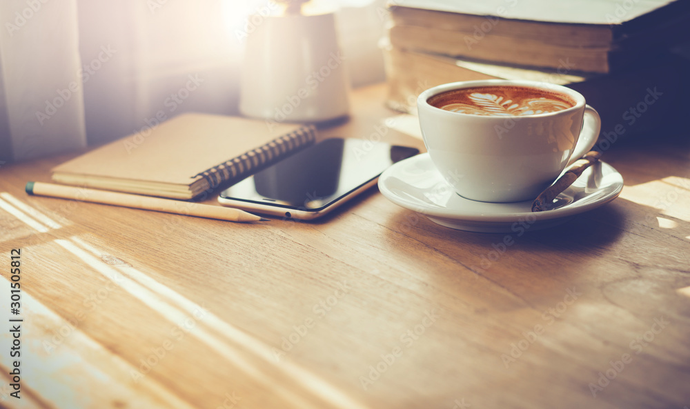 Closeup latte art coffee in white cup with smartphone and notebook on wooden table with sunlight through the window. Vintage color tone Closeup latte art coffee in white cup with smartphone and notebook on wooden table with sunlight through the window. Vintage color tone