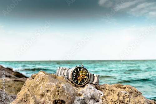 an exotic stainless big dial silver watch kept on brownish rocks at the beach and greenish sea water in the background and clouds on the sky