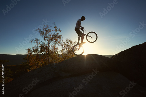 Silhouette of professional cyclist standing on back wheel on trial bicycle. Sportsman rider balancing on the edge of big boulder on the top of mountain at sunset. Concept of extreme sport