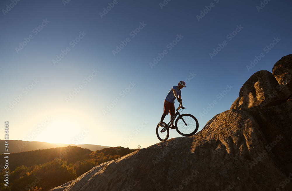 Silhouette of professional bicyclist balancing on trial bicycle on top of big boulder, male rider making acrobatic stunt on summer evening, blue sky and sunset on background. Concept of extreme sport Silhouette of professional bicyclist balancing on trial bicycle on top of big boulder, male rider making acrobatic stunt on summer evening, blue sky and sunset on background. Concept of extreme sport