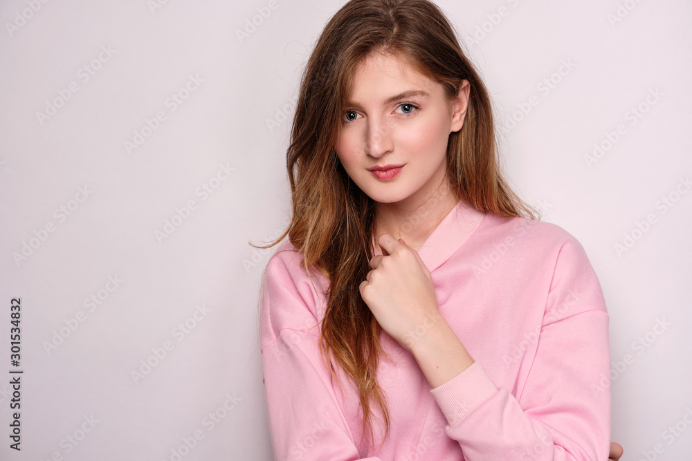 A girl in a pink sweatshirt on a light background straightens the gate and looks into the frame. A girl in a pink sweatshirt on a light background straightens the gate and looks into the frame.