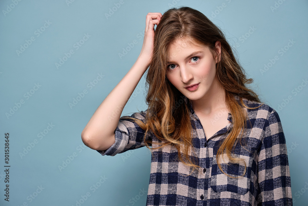 A blue-eyed girl in a plaid shirt stands on a blue background and looks into the frame, holding her hand to her head A blue-eyed girl in a plaid shirt stands on a blue background and looks into the frame, holding her hand to her head