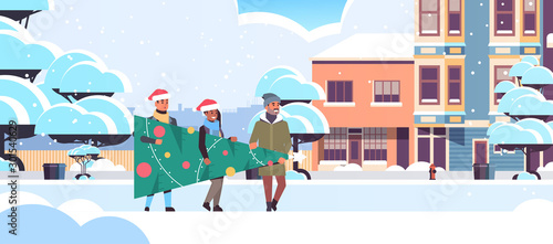 people carrying fir tree preparing for merry christmas happy new year holiday celebration concept mix race friends wearing santa hats snowy city street cityscape background horizontal full length