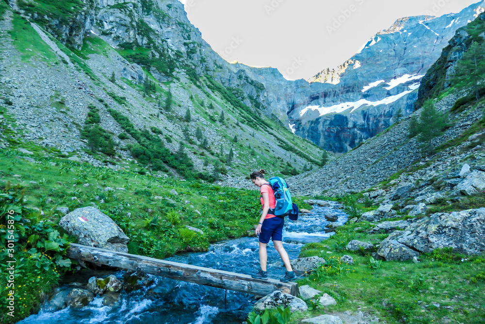 Young woman with a backpack crosses the small river, flowing between tall mountain peaks. Some of the slopes are covered with snow. In the back is another mountain range. Spring in alpine valleys