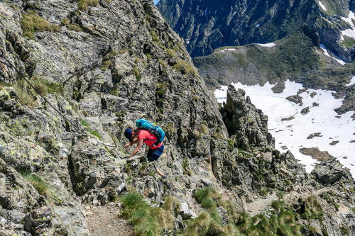 A young woman with a big backpack hikes down a steep slope using a metal rope. Girl is focused and concentrated. Dangerous hike. Steep via ferrata.There is a lot of snow in the valley below. photo