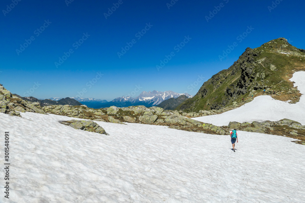 A young woman with a big backpack hikes through a snowy plateau in high mountains. Girl is wearing shorts.In the back there is a sharp mountain peak. Schladming Alps in Austria. Outdoor activities.