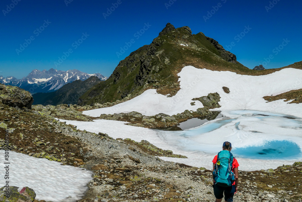 Obraz premium Young woman enjoying the view a frozen alpine lake. The ice shines in many different shades of blue. Lake is surrounded by tall mountains. No snow on the peaks Spring slowly coming to high altitudes