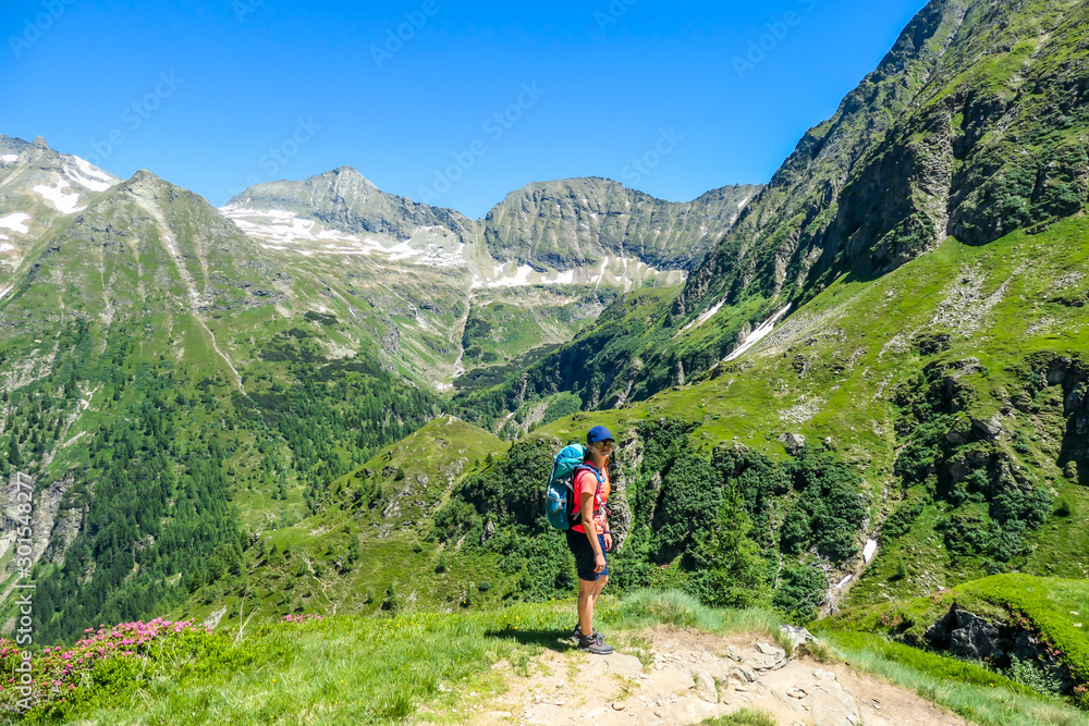 A young woman with a big backpack hikes down on a steep pathway between tall mountain peaks. Some of the slopes are covered with snow. In the back is another mountain range. Spring in alpine valleys.