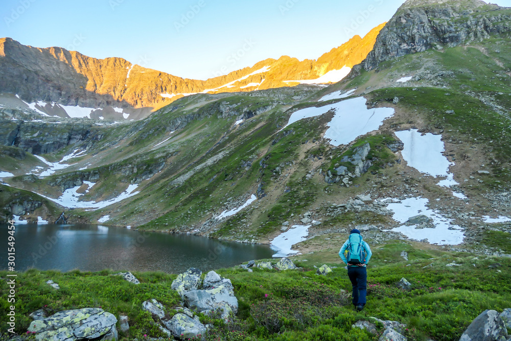 A young woman with a big hiking backpack standing next to a clear, navy blue lake hiding between tall mountain peaks. Some slopes are covered with snow. Soft colours of sunrise over the high peaks