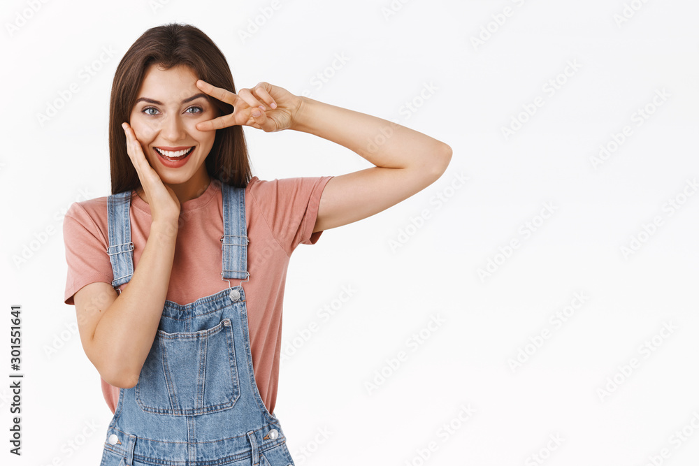 Sassy, cheerful young woman in overalls, t-shirt, showing peace or victory sign over eye in disco motion, touching cheek gently, look determined and confident, standing white background self-assured Sassy, cheerful young woman in overalls, t-shirt, showing peace or victory sign over eye in disco motion, touching cheek gently, look determined and confident, standing white background self-assured