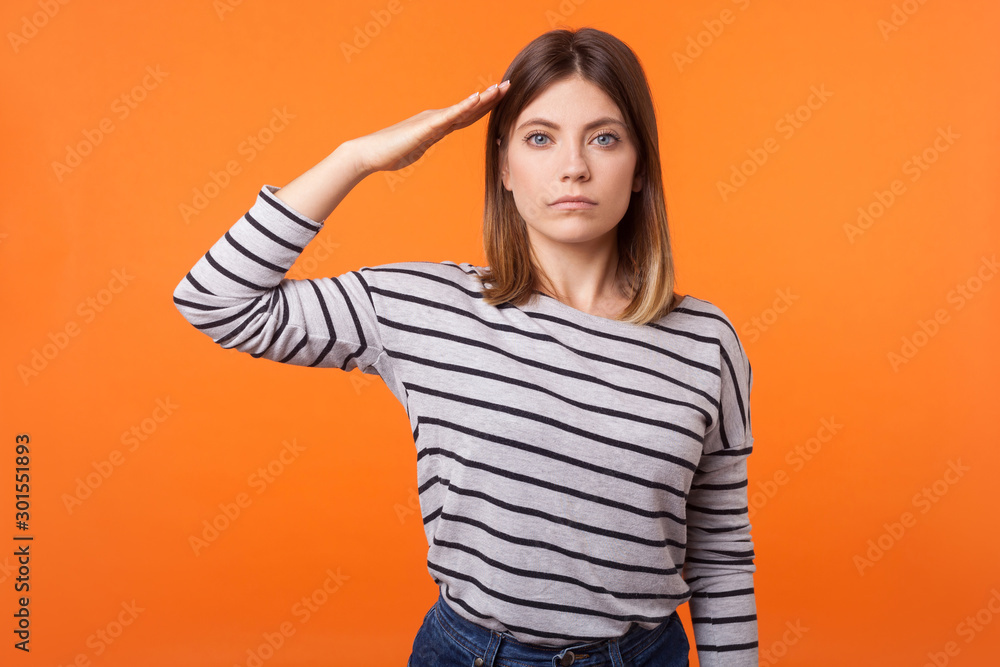Yes sir! Portrait of obedient serious young woman with brown hair in long sleeve striped shirt standing, making military salute with hand near head. indoor studio shot isolated on orange background Yes sir! Portrait of obedient serious young woman with brown hair in long sleeve striped shirt standing, making military salute with hand near head. indoor studio shot isolated on orange background