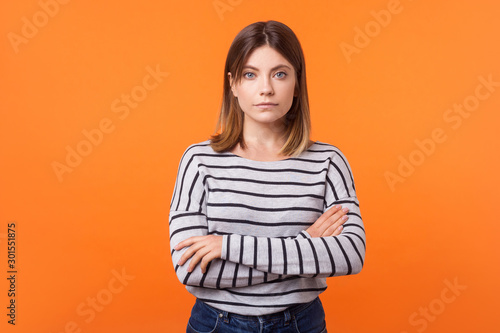 Portrait of focused serious young woman with brown hair in long sleeve striped shirt standing with crossed arms and looking attentively at camera. indoor studio shot isolated on orange background