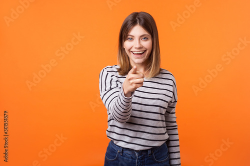 Hey you! Portrait of adorable positive woman with brown hair in long sleeve striped shirt standing, smiling happily and pointing finger at camera. indoor studio shot isolated on orange background