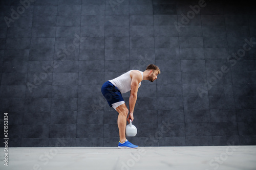 Strong muscular handsome Caucasian man in shorts and t-shirt standing outdoors and lifting kettle bell. In background is gray wall.