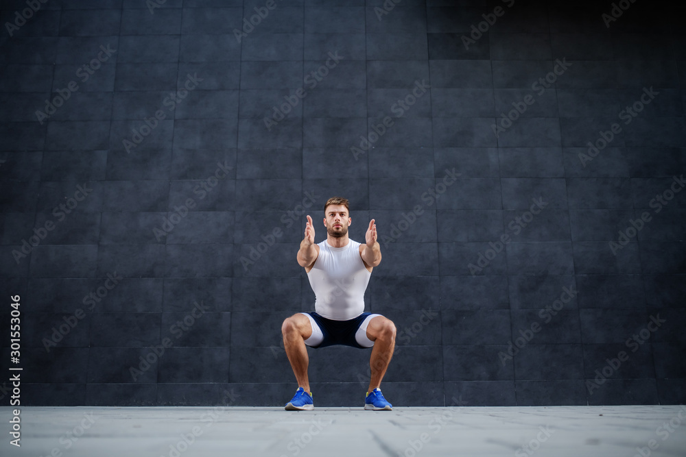 Handsome muscular caucasian man in shorts and t-shirt doing squatting exercise outdoors. In background is gray wall.