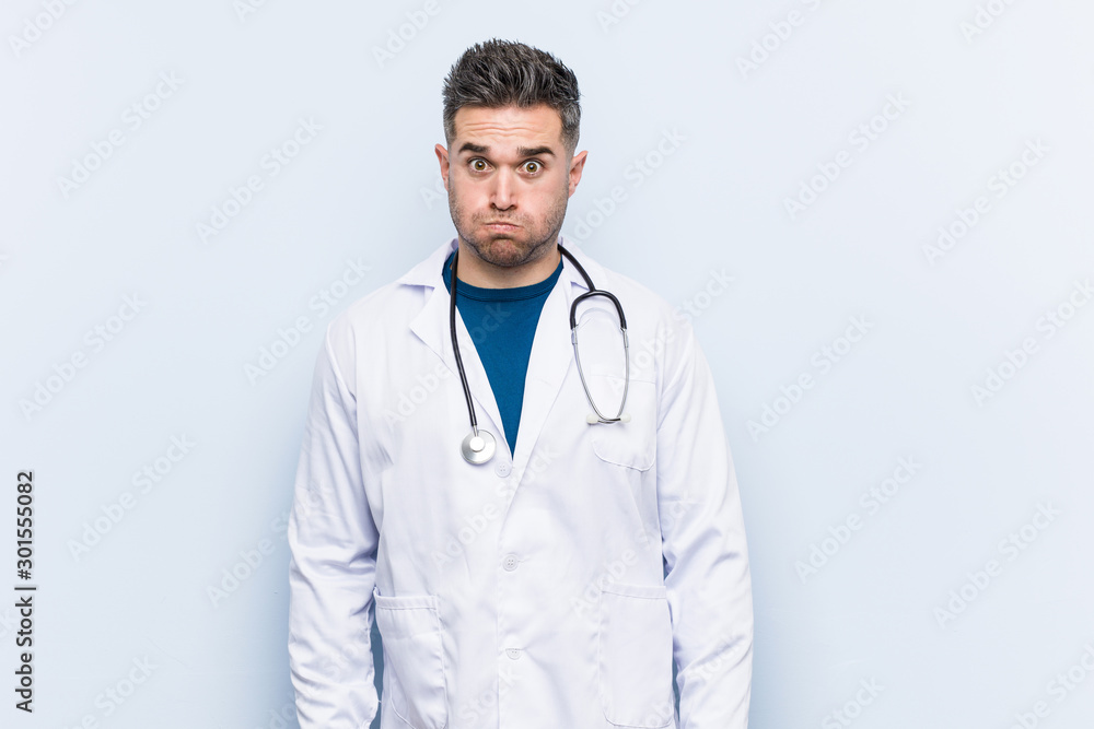 Young handsome doctor man blows cheeks, has tired expression. Facial expression concept.