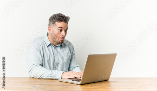 Young handsome man working with his laptop shrugs shoulders and open eyes confused.