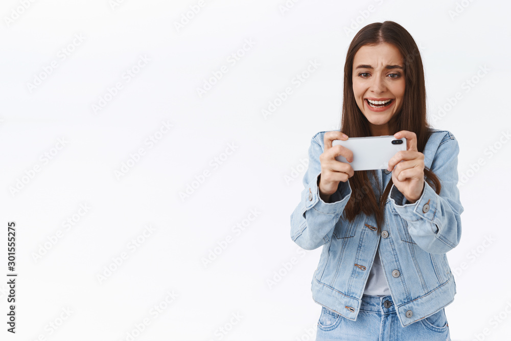 Distressed, intense young attractive woman in denim, holding smartphone horizontally, frowning and grimacing as losing online mobile game battle, playing on telephone, standing white background Distressed, intense young attractive woman in denim, holding smartphone horizontally, frowning and grimacing as losing online mobile game battle, playing on telephone, standing white background