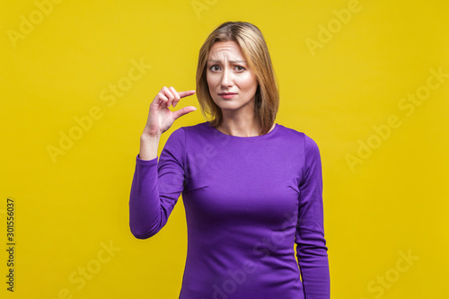 Portrait of disappointed woman in elegant purple dress frowning and showing a little bit gesture, asking some more, dissatisfied upset with amount. indoor studio shot isolated on yellow background
