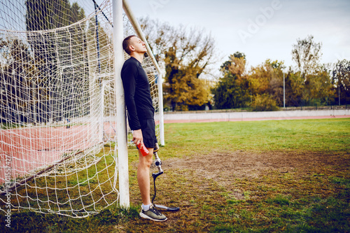 Side view of handsome sporty handicapped man in sportswear and with artificial leg while standing on football court and holding refreshment. photo