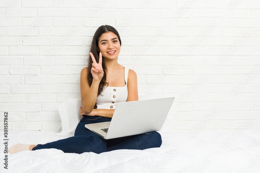 Young arab woman working with her laptop on the bed joyful and carefree showing a peace symbol with fingers.