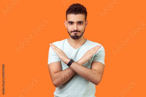 No way, absolutely not. Portrait of determined brunette man with beard in white t-shirt showing x sign with crossed hands, meaning stop, this is the end. studio shot isolated on orange background