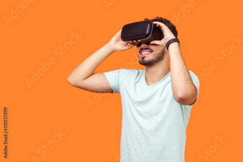 Portrait of happy gamer, brunette man with beard in t-shirt wearing vr headset, playing virtual reality game with smile on his face, innovative technology. studio shot isolated on orange background