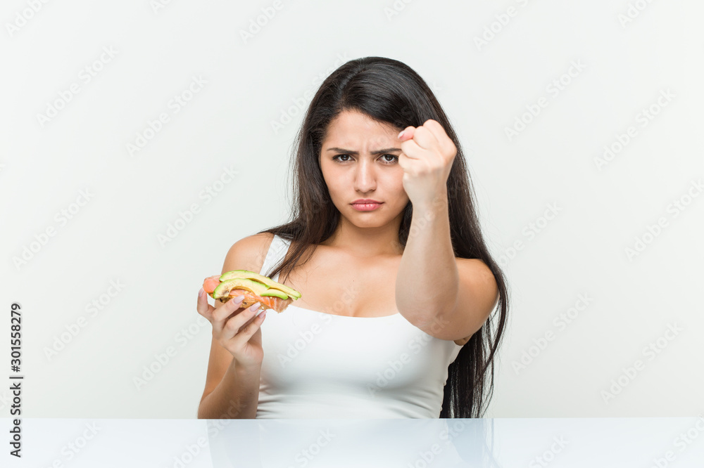 Young hispanic woman holding an avocado toast showing fist to camera, aggressive facial expression. Young hispanic woman holding an avocado toast showing fist to camera, aggressive facial expression.