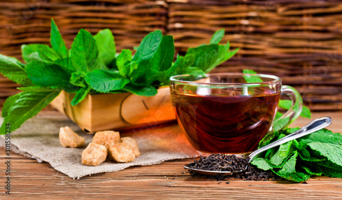 Cup of black tea with dried tea in spoon, brown sugar and fresh mint leaves in basket on a wooden background.