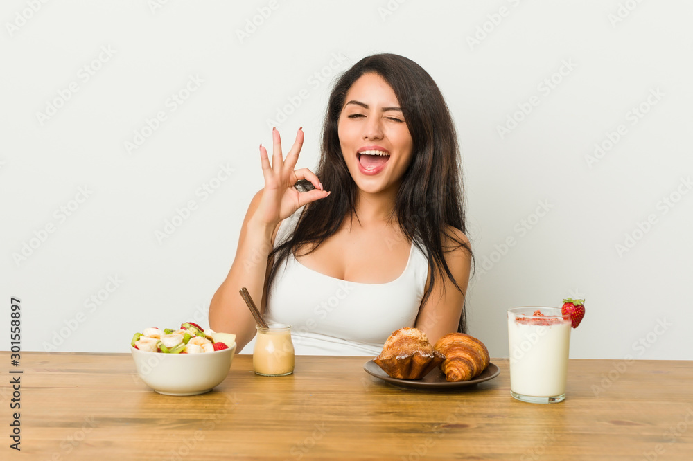 Young curvy woman taking a breakfast winks an eye and holds an okay gesture with hand. Young curvy woman taking a breakfast winks an eye and holds an okay gesture with hand.