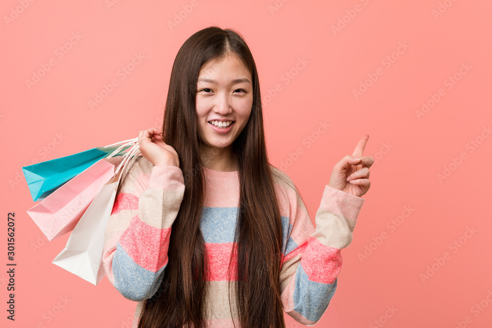 Young asian woman holding a shopping bag smiling cheerfully pointing with forefinger away.
