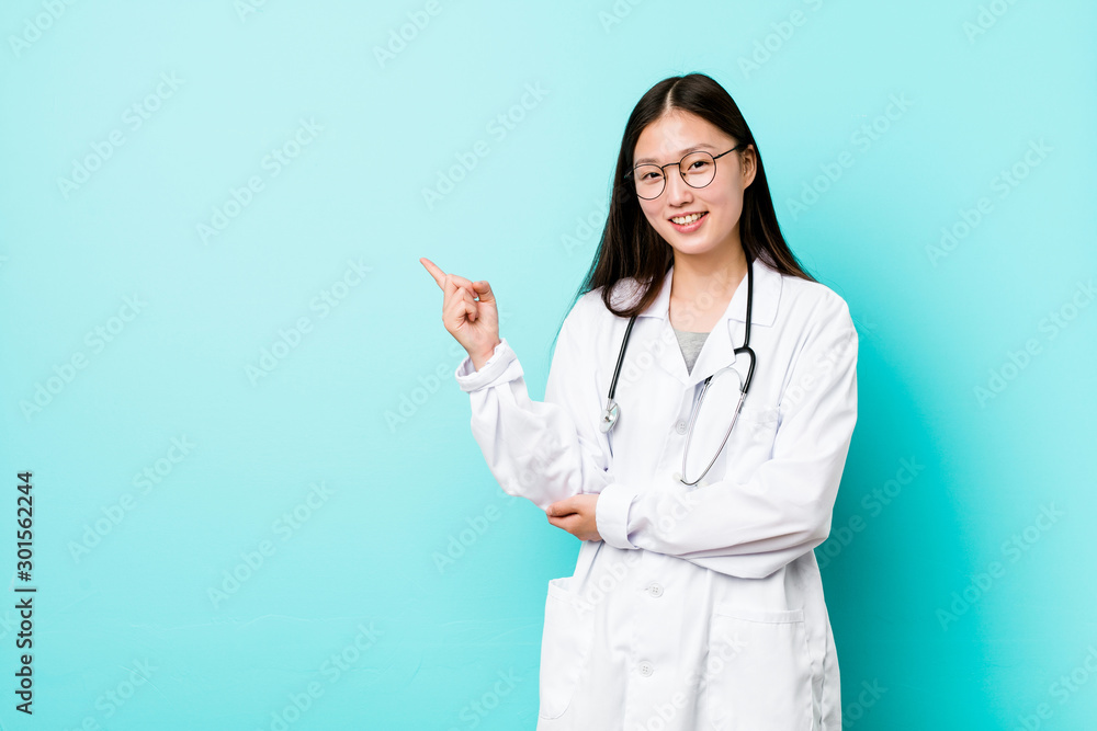 Young chinese doctor woman smiling cheerfully pointing with forefinger away. - obrazy, fototapety, plakaty 