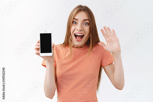 Amused young caucasian woman in striped t-shirt looking upbeat, hold smartphone, showing black mobile display, waving one hand, say bye to ex-boyfriend as unfollow his profile, white background photo