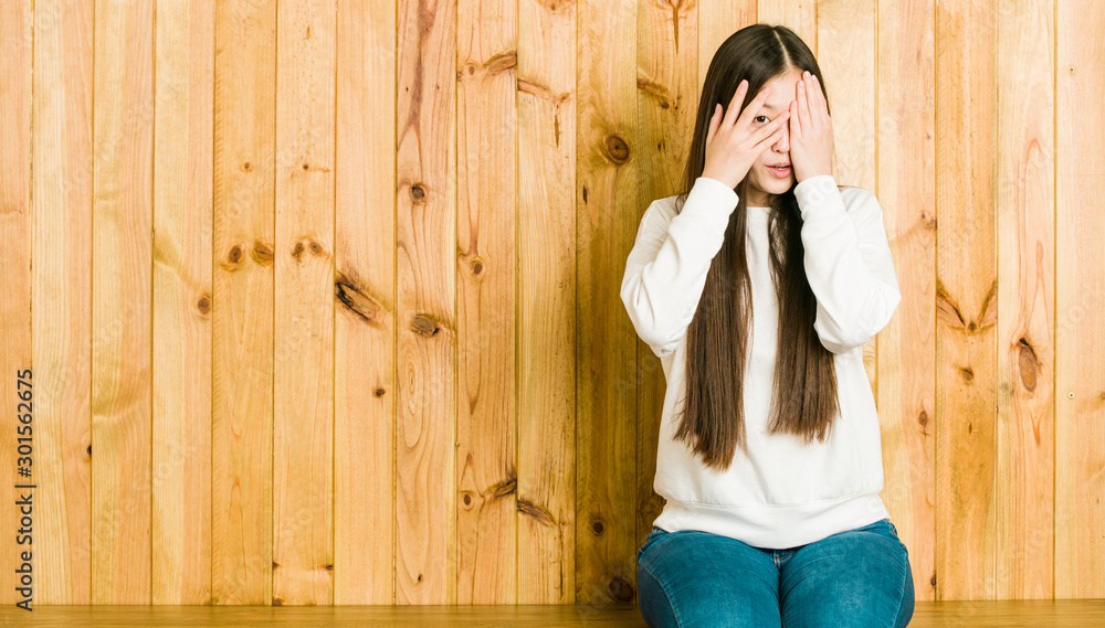 Obraz premium Young chinese woman sitting on a wooden place blink through fingers frightened and nervous.