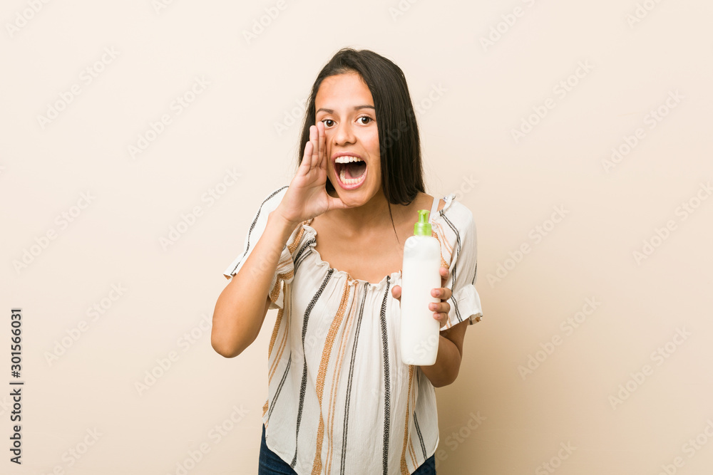 Young hispanic woman holding a cream bottle shouting excited to front. Young hispanic woman holding a cream bottle shouting excited to front.