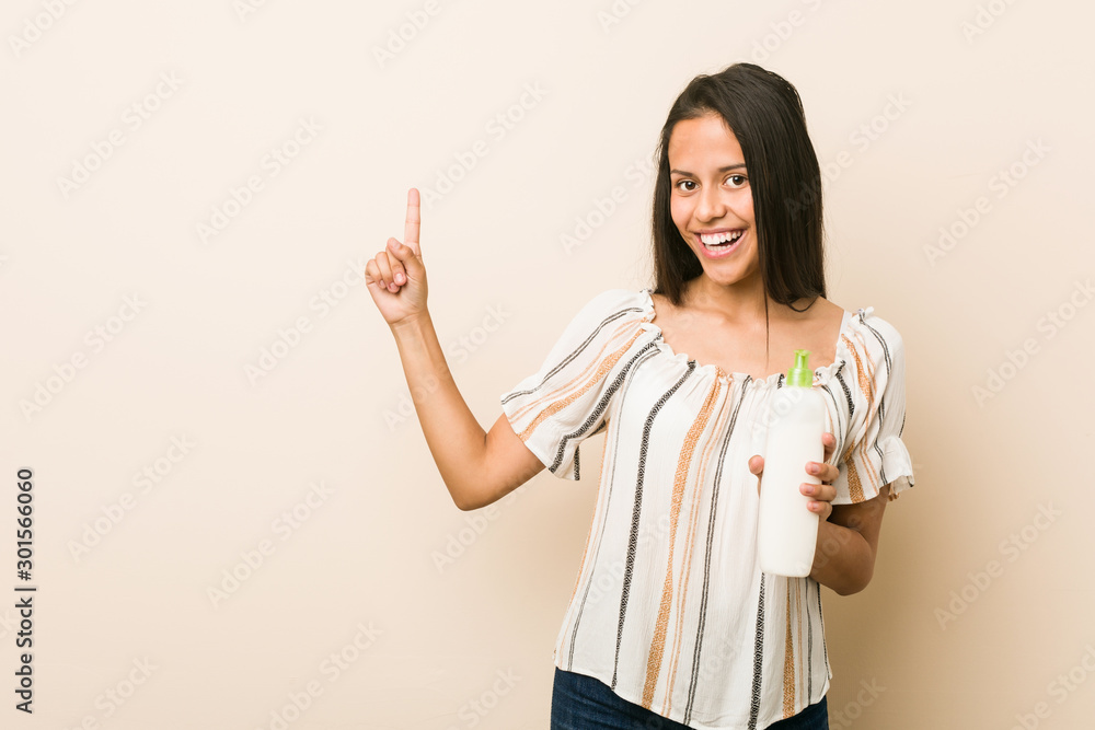 Young hispanic woman holding a cream bottle smiling cheerfully pointing with forefinger away. Young hispanic woman holding a cream bottle smiling cheerfully pointing with forefinger away.