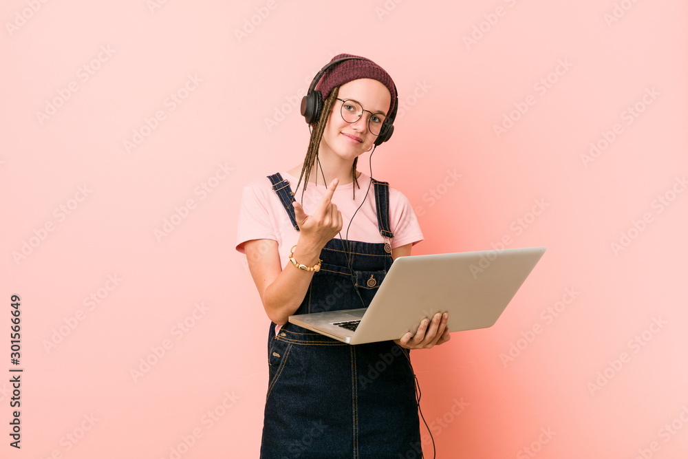 Young caucasian woman holding a laptop pointing with finger at you as if inviting come closer. Young caucasian woman holding a laptop pointing with finger at you as if inviting come closer.